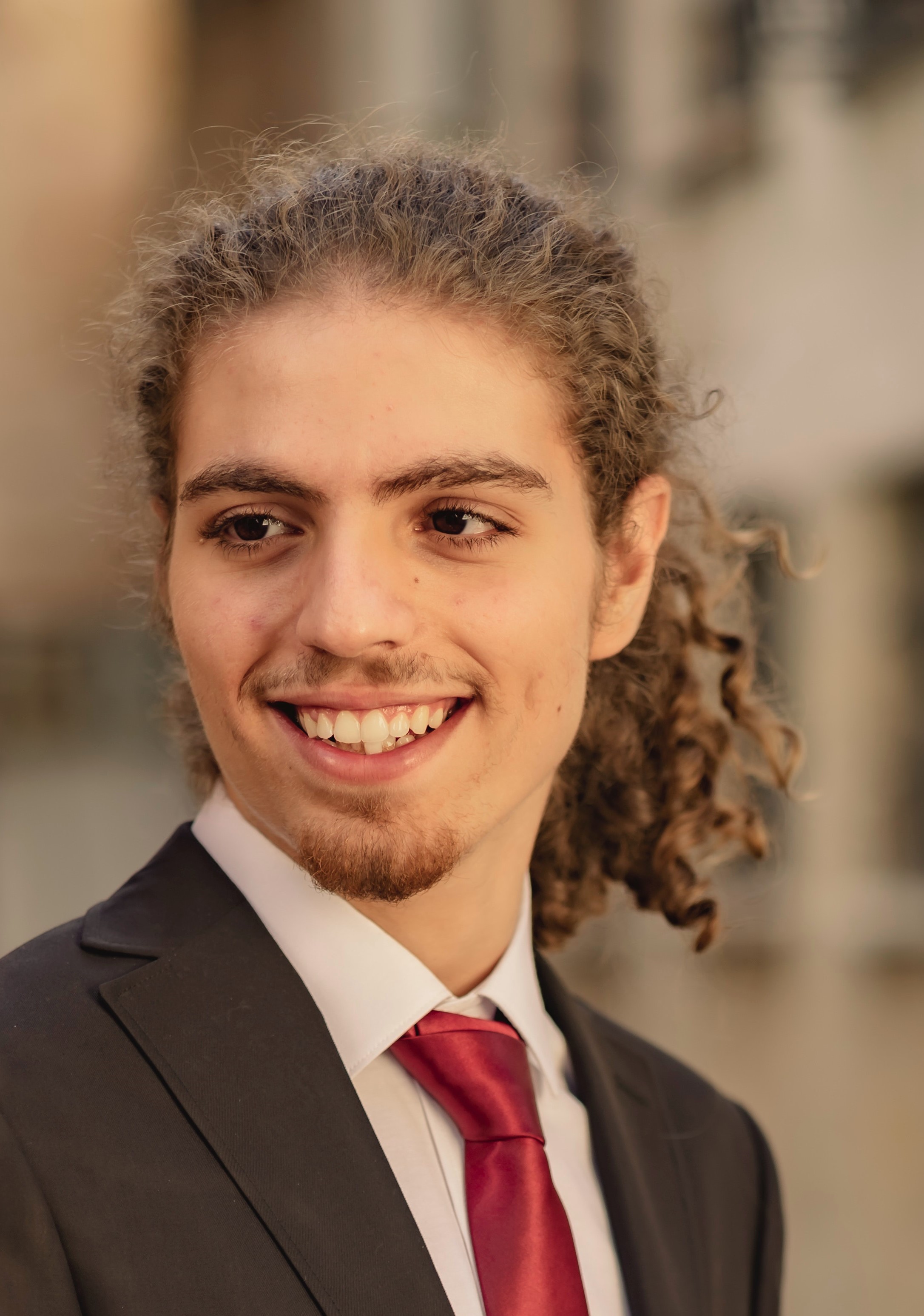 Man in suit with tie smiling with long curly hair tied back and a small amount of facial hair