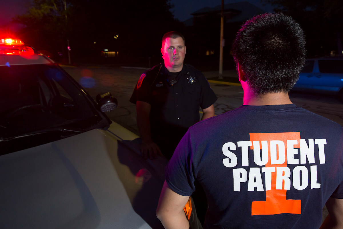a police officer faces a student wearing an Illinois Tshirt which reads "student patrol", with a police car off to the side
