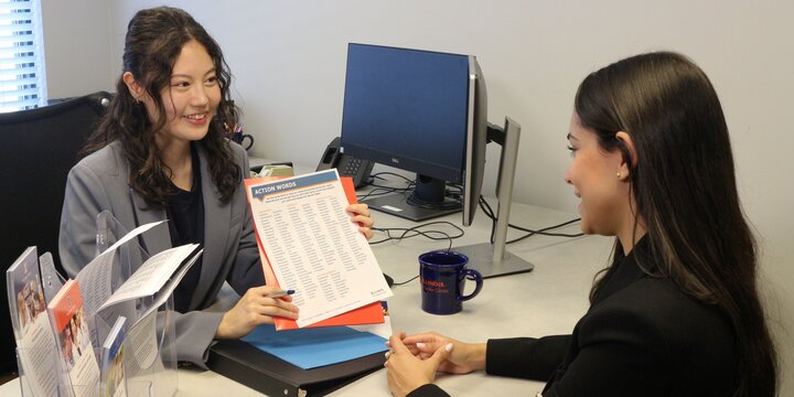 A graduate assistant shows a student sitting at a desk a flyer labeled "Action Words"