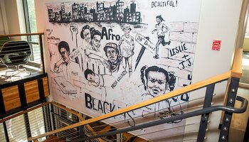 Black and white mural painted on interior wall in stairwell of BNAAC, with images of African Americans and a city, and phrases like "Black is...", "Afro," "Beautiful," and more.