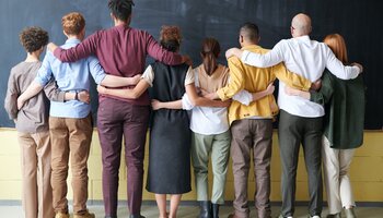 A group of 8 people of varying heights, genders, ages and races standing with their backs to the camera, facing a chalkboard on the wall, with arms around eachother's shoulders and waists